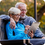 An older man and older woman sit on a bench together and peer at a tablet.